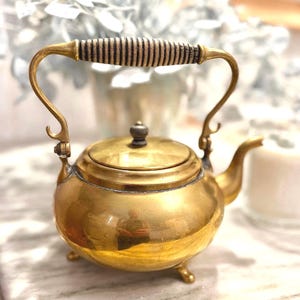 May include: A small, antique brass teapot with a wooden handle. The teapot has a rounded body, a curved spout, and three small feet. The handle is made of a dark wood with a ridged design. The teapot is highly polished and reflects the surrounding light.