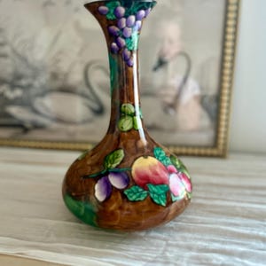 May include: A ceramic vase with a bulbous base and a slender neck, featuring a glossy brown glaze decorated with hand-painted clusters of purple grapes, green leaves, and red and yellow apples arranged around the exterior surface.