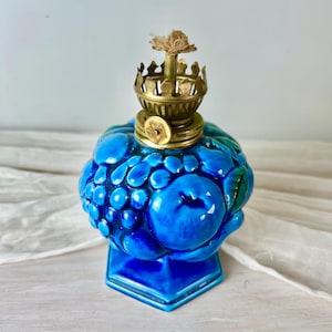 Könnte beinhalten: Eine leuchtend blaue Öllampe aus Glas mit einem geformten Frucht- und Traubenmotiv. Die Lampe verfügt über einen Messingbrenner mit einem zentralen Docht und einem kleinen runden Einstellknopf an der Vorderseite. Sie steht auf einem sechseckigen Sockel.