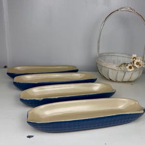 May include: Four elongated, corn-shaped serving dishes with a smooth, cream-colored interior and a textured, deep blue exterior. Each dish features a realistic kernel pattern on the outside, arranged in a row, accompanied by a small white basket.