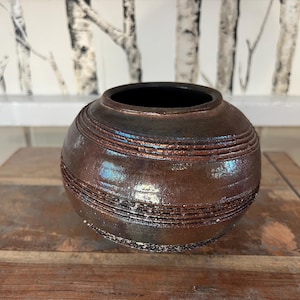 May include: A dark brown ceramic vase with a textured, iridescent finish. The vase has a round shape and a wide opening.