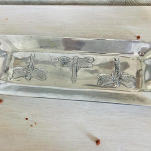 Könnte beinhalten: Ein rechteckiges, silberfarbenes Tablett mit drei geprägten Libellenmotiven. Das Tablett hat eine leicht gebogene Form mit erhöhten Kanten und Griffen an jedem Ende. Die Libellen-Details verleihen diesem Servierstück eine dekorative Note.