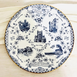 May include: A round white ceramic decorative plate with a gold rim, featuring blue ink illustrations of Bath landmarks including Bath Abbey, Pulteney Bridge, and Royal Crescent. The text reads BATH and WORLD HERITAGE CITY with floral accents.