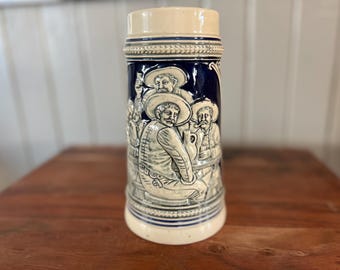 May include: A white ceramic beer stein with a blue and white design. The stein features a scene of four men in traditional clothing, drinking and smoking.