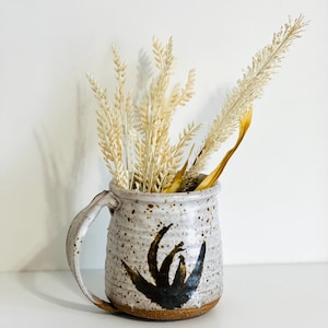 Puede incluir: Una taza de cerámica blanca con asa marrón y un diseño abstracto negro. La taza está llena de hierbas secas blancas y marrones.