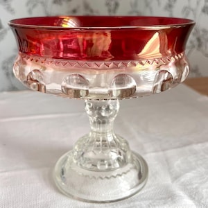 May include: A clear glass pedestal bowl with a vibrant red rim. The bowl features a decorative pattern around the base and a detailed stem. This elegant piece is perfect for serving desserts or displaying decorative items.