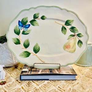 May include: An off-white ceramic platter with a scalloped edge. The platter has a painted design of green leaves, a blue butterfly, and a pear. The platter is supported by a metal stand and rests on a book.