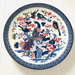 May include: A white ceramic plate with a blue and white border. The centre of the plate features a floral design with red, blue, and yellow flowers. There are also several blue and black birds with long tails.