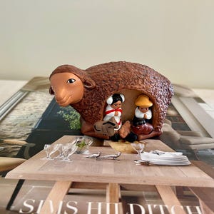 May include: A brown ceramic sheep figurine with a hollowed-out center, housing two small figures in traditional attire. The sheep is placed on a table setting with glassware and plates. The figurine is a unique decorative item.