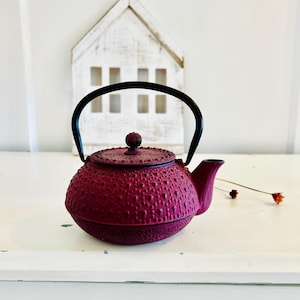 Peut inclure: Une théière en fonte magenta foncé texturée avec une poignée et un bec noirs. La théière a une forme arrondie et un couvercle assorti avec un petit bouton. Une maison en bois décorative est en arrière-plan.