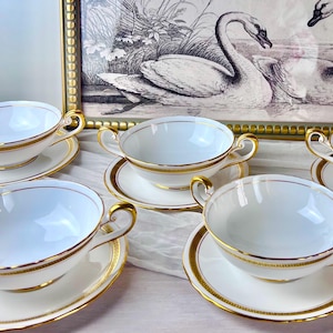 May include: A set of white porcelain bouillon cups and saucers featuring two handles on each cup. The set is decorated with a gold Greek key pattern along the rims and bases, complemented by thin gold trim on the edges and handles.