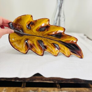 May include: A ceramic leaf-shaped dish with a glossy finish. The dish features a blend of amber, brown, and black hues, creating a marbled effect. The leaf design is detailed with textured edges and a smooth, curved surface. A decorative piece for serving or display.