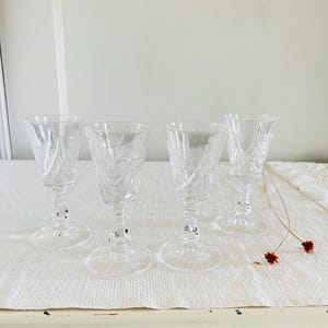 May include: Four clear crystal cordial glasses with etched floral patterns. The glasses have a stemmed base and a flared bowl. They are arranged on a white surface, with a neutral background.