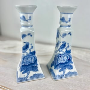 May include: A pair of white porcelain candlesticks featuring a blue hand-painted pattern of birds perched on branches above large floral blossoms. Each candlestick has a flared base, a tapered body, and a wide rim at the top for candles.
