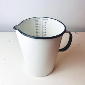 May include: White enamel measuring jug with a black rim and handle. The jug has a spout and measurement markings in ounces and millilitres. A vintage kitchenware item.