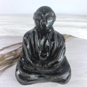May include: A small, hand-sculpted ceramic figurine of a seated figure in a meditative pose. The sculpture features a glossy, deep black glaze with subtle greyish-white highlights along the textured folds of the clothing and facial features.