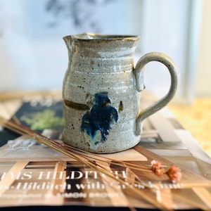 May include: A small, handmade ceramic jug with a light grey glaze and blue and brown accents. The jug has a rounded body and a handle.