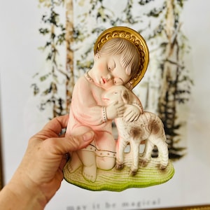 May include: A vintage figurine of a child holding a lamb. The child is wearing a pink robe and a gold halo. The figurine is made of ceramic and has a painted finish.