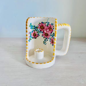 Puede incluir: Un portavelas de cerámica blanca con forma de taza y asa. El interior presenta flores rosas pintadas a mano con enredaderas verdes. Los bordes de la taza y el soporte para la vela están decorados con un patrón de cuadros amarillos y blancos.