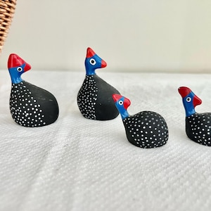 May include: Four decorative guinea fowl figurines. Each bird is black with white polka dots, a red head, and a blue neck. The figurines are arranged on a white surface near a wicker basket.