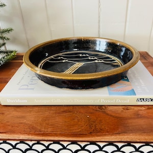 May include: A black ceramic dish with a gold rim and a white wave design. The dish is sitting on a book titled "Antique Collector's Directory of Period Detail" by Davidson.