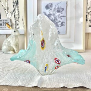 May include: A clear glass decorative bowl with an unusual, organic form. The bowl has a white speckled interior with colourful accents of red, yellow, and blue. The rim has a light blue hue.