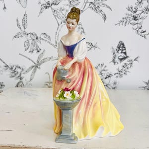 May include: A porcelain figurine of a woman in a long, flowing gown, holding a watering can. The dress is a gradient of yellow, pink, and orange. A small pedestal holds a flower arrangement. The figurine is approximately 8 inches tall.