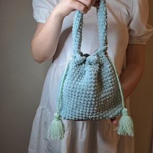 Peut inclure: Un sac à cordon en crochet bleu clair avec des glands verts. Le sac a une texture moelleuse et est tenu par la main d'une personne.