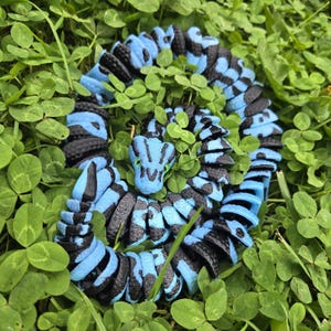 Pu&ograve; includere: Un giocattolo a forma di serpente blu e nero arrotolato, con un motivo dettagliato, appoggiato in un letto di trifogli verdi. La testa del serpente &egrave; visibile, con occhi verdi e un design dettagliato. Il giocattolo &egrave; realizzato in un materiale resistente.