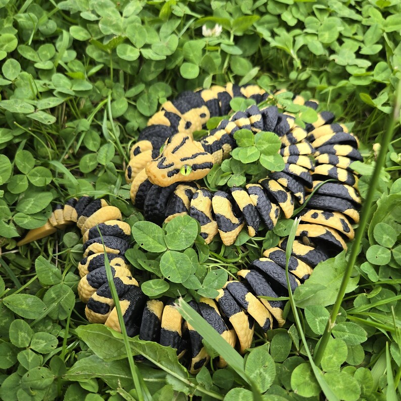 Pu&ograve; includere: Una statuetta di serpente giallo e nero arrotolata, con occhi verdi, riposa tra trifogli ed erba verdi. Il corpo del serpente &egrave; segmentato con un motivo ripetuto di nero e giallo. La testa &egrave; dettagliata con squame strutturate.
