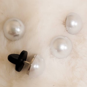 May include: Four white pearl buttons, one with a black plastic post and metal backing. The buttons are on a white fluffy surface.