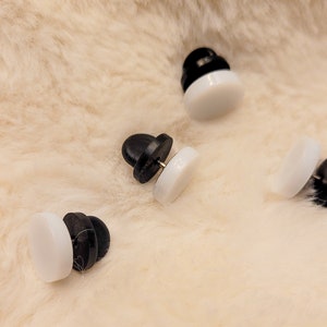 May include: Four black and white enamel pin backs. The pin backs are round and have a flat back. The pin backs are used to attach pins to clothing or other items.