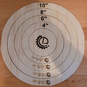 Puede incluir: Un blanco de madera con círculos concéntricos marcados a 10 cm, 15 cm, 20 cm y 25 cm. El centro presenta un logotipo con el texto "Clark Creative design". Ideal para juegos o decoración.