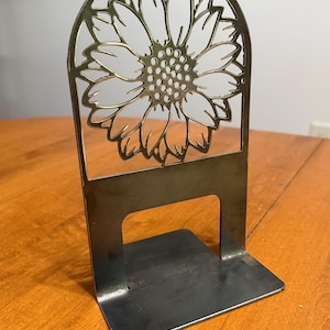 Può includere: Un fermalibri in metallo con un design a girasole. Il girasole è ritagliato dal metallo, creando un pezzo decorativo e funzionale. Il fermalibri è di colore argento e ha una base robusta.