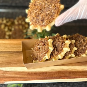 May include: A round, scalloped-edge pastry topped with a layer of toasted, shredded brown coconut. The pastry has a light-colored, baked dough base. Several identical pastries are arranged in a rectangular cardboard tray, with one held up.