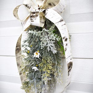 May include: A decorative hanging arrangement featuring a large birch bark ribbon bow. The arrangement includes green and gray foliage, small white flowers, and yellow accents. The background is a white, textured surface.