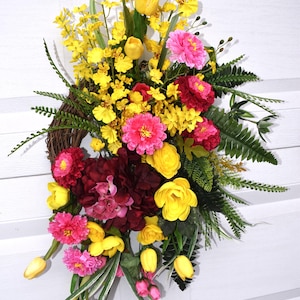 May include: A spring-themed wreath with yellow tulips, pink and red flowers, and green ferns. The wreath is made of grapevine and is decorated with a variety of artificial flowers.