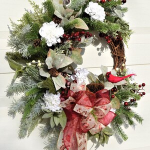 May include: A Christmas wreath with a red cardinal, white hydrangeas, red berries, pine branches, and a red and gold bow.