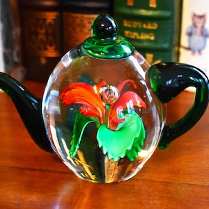 May include: A clear glass teapot with a green handle, spout, and lid. Inside the teapot is a colorful floral design with red, orange, and green elements. The teapot is sitting on a wooden surface.