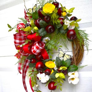 May include: A decorative wreath featuring red apples, yellow flowers, and white blossoms. The wreath is adorned with green leaves, butterflies, and a red and white checkered bow. The wreath is made of woven twigs.