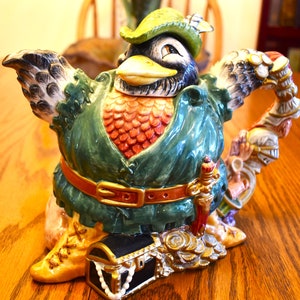 May include: A ceramic teapot shaped like a bird wearing a green and gold outfit, holding a sword and a treasure chest. The bird has a black beak and brown eyes.
