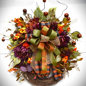 May include: A fall-themed wreath with a brown grapevine base, decorated with burgundy and orange flowers, green and orange plaid ribbon, and brown berries.
