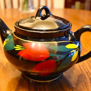 May include: Black teapot with a floral design in red, yellow, and green. The teapot has a lid and a handle.