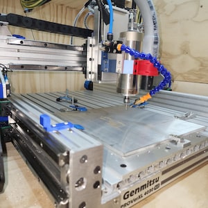 May include: A CNC machine with a silver and blue frame, featuring a cutting head with red and blue cooling tubes. The machine has a metal work surface and the text "Genmitsu PROVerXL 4030" is visible.