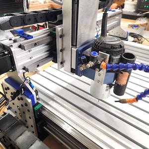 May include: A CNC machine with a silver frame and a black motor. The machine has blue and black hoses and wires. The work surface has parallel grooves. The machine is designed for precision cutting and fabrication.