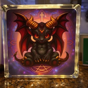 Puede incluir: Un bloque de vidrio decorativo con un demonio de dibujos animados con ojos, cuernos y alas rojas. La obra de arte está ambientada sobre un fondo morado y naranja con una cruz y un pentagrama. El bloque está iluminado.