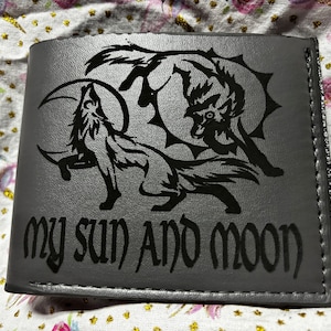 Puede incluir: Cartera gris plegable con un diseño de lobo negro y el texto "my sun AND moon". El diseño presenta dos lobos, uno aullando a la luna y el otro frente a un sol. La cartera está hecha de un material texturizado.