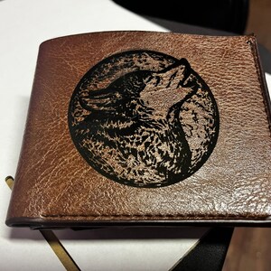 May include: A brown leather wallet with a black wolf silhouette design. The wolf is howling at the moon.