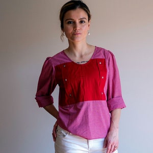 Puede incluir: Una mujer con una camisa de cuadros rojos y rosas con un panel rojo en la parte delantera. La camisa tiene cierre de botones y cuello. Lleva pantalones blancos.