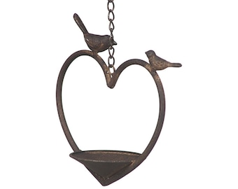 Op de afbeelding: Een rustieke metalen vogelvoederhuisje in de vorm van een hart met twee kleine vogels die bovenop zitten. De voederbak heeft een klein kommetje aan de onderkant voor vogelzaad.